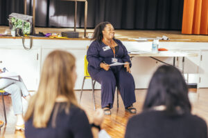 Facilitator leading a workshop, sitting on a yellow chair with two students in front