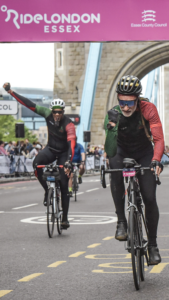 Paul and Adisa at the finish line of ride London, Adisa in the background with a fist raised and Paul in the foreground fist pumped.